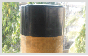 A black heat-shrink cap recovered over the sawn top of a round timber pole outdoors, its lower edge sitting tight and straight against the timber, with trees behind